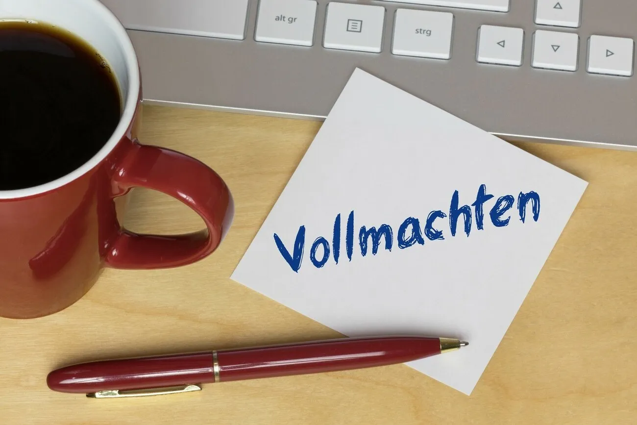 Ein Zettel mit der Aufschrift "Vollmachten" liegt halb auf einer Tastatur. Daneben steht eine rote Kaffeetasse und darunter ein roter Kugelschreiber.